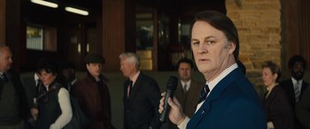 Movie still from “Eddie the Eagle” (2015), directed by Dexter Fletcher – A man holding a microphone while standing next to other men; Medium shot, Over the shoulder angle