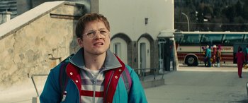 Movie still from “Eddie the Eagle” (2015), directed by Dexter Fletcher – A young man wearing glasses standing on the sidewalk; Close Up shot, Low angle