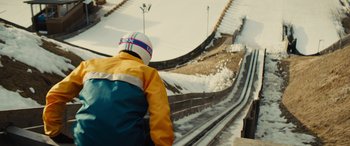 Movie still from “Eddie the Eagle” (2015), directed by Dexter Fletcher – A man riding a snowboard down the side of a ski slope; Wide shot, Low angle