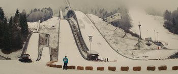 Movie still from “Eddie the Eagle” (2015), directed by Dexter Fletcher – A person standing on a hill with a ski slope in the background; Extreme Wide shot, Low angle