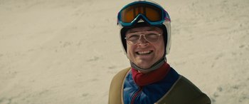 Movie still from “Eddie the Eagle” (2015), directed by Dexter Fletcher – A man wearing a helmet and goggles smiles for the camera; Close Up shot, Over the shoulder angle
