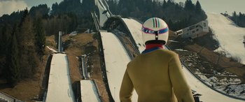 Movie still from “Eddie the Eagle” (2015), directed by Dexter Fletcher – A person wearing a helmet standing on top of a ski slope; Wide shot, Low angle