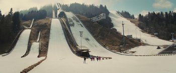 Movie still from “Eddie the Eagle” (2015), directed by Dexter Fletcher – A group of skiers are standing in the snow near a ski jump; Extreme Wide shot, High angle