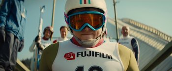 Movie still from “Eddie the Eagle” (2015), directed by Dexter Fletcher – A person wearing a helmet and goggles in front of a group of people; Close Up shot, Low angle