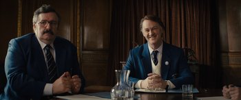 Movie still from “Eddie the Eagle” (2015), directed by Dexter Fletcher – A man sitting at a table with another man in front of him; Medium shot, Over the shoulder angle