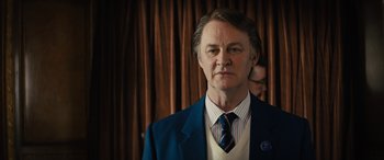 Movie still from “Eddie the Eagle” (2015), directed by Dexter Fletcher – A man in a suit and tie standing in front of a curtain; Close Up shot, Over the shoulder angle