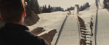 Movie still from “Eddie the Eagle” (2015), directed by Dexter Fletcher – A person riding a snowboard down a snow covered slope; Extreme Wide shot, Over the shoulder angle