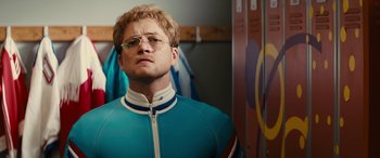 Movie still from “Eddie the Eagle” (2015), directed by Dexter Fletcher – A man in a blue and white suit with glasses; Close Up shot, Over the shoulder angle