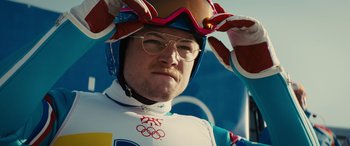 Movie still from “Eddie the Eagle” (2015), directed by Dexter Fletcher – A man wearing a helmet and glasses while standing on a ski slope; Close Up shot, Low angle