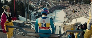 Movie still from “Eddie the Eagle” (2015), directed by Dexter Fletcher – A man in blue and white ski suit on a ski slope; Wide shot, High angle