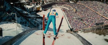 Movie still from “Eddie the Eagle” (2015), directed by Dexter Fletcher – A person skiing down a ski slope with a crowd watching; Extreme Wide shot, Overhead angle