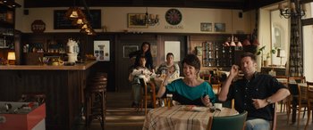 Movie still from “Eddie the Eagle” (2015), directed by Dexter Fletcher – A group of people sitting at a table in a restaurant; Medium shot, Over the shoulder angle