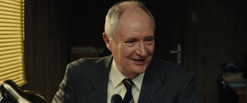 Movie still from “Eddie the Eagle” (2015), directed by Dexter Fletcher – An older man wearing a suit and tie while talking into a microphone; Close Up shot, High angle
