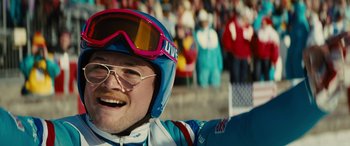 Movie still from “Eddie the Eagle” (2015), directed by Dexter Fletcher – A man wearing a helmet and goggles smiles for the camera; Close Up shot, Over the shoulder angle
