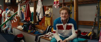 Movie still from “Eddie the Eagle” (2015), directed by Dexter Fletcher – A man sitting on top of a bench in a room; Medium shot, Over the shoulder angle