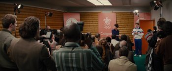 Movie still from “Eddie the Eagle” (2015), directed by Dexter Fletcher – A crowd of people gathered in front of a microphone; Wide shot, Over the shoulder angle