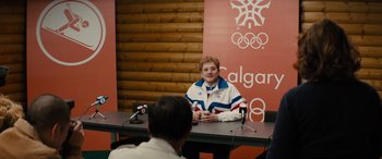 Movie still from “Eddie the Eagle” (2015), directed by Dexter Fletcher – A man sitting at a table in front of microphones; Medium shot, Over the shoulder angle