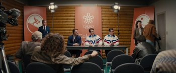 Movie still from “Eddie the Eagle” (2015), directed by Dexter Fletcher – Three men are sitting at a table in front of a woman; Wide shot, Over the shoulder angle
