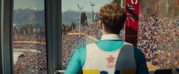 Movie still from “Eddie the Eagle” (2015), directed by Dexter Fletcher – A man sitting in front of a large crowd of people; Medium shot, Over the shoulder angle