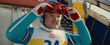 Movie still from “Eddie the Eagle” (2015), directed by Dexter Fletcher – A man wearing a helmet and goggles while standing in the snow; Close Up shot, Over the shoulder angle