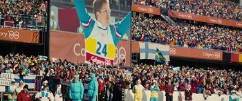 Movie still from “Eddie the Eagle” (2015), directed by Dexter Fletcher – A crowd of people standing in front of a large picture of a man; Extreme Wide shot, Over the shoulder angle