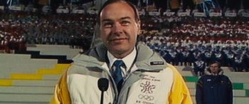 Movie still from “Eddie the Eagle” (2015), directed by Dexter Fletcher – A man wearing a jacket and tie standing in front of a microphone; Close Up shot, Low angle