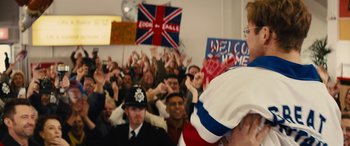Movie still from “Eddie the Eagle” (2015), directed by Dexter Fletcher – A group of people in a room waving flags and holding signs; Medium shot, Over the shoulder angle