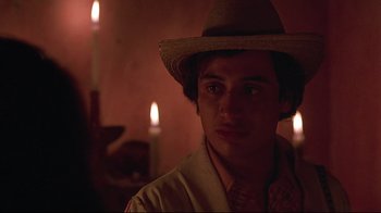 Movie still from “El Norte” (1983), directed by Gregory Nava – A young man wearing a straw hat in front of candles in a dimly - lit room; Close Up shot, Over the shoulder angle