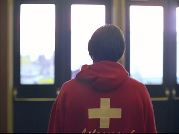 Movie still from “Elephant” (2003), directed by Gus Van Sant – A person wearing a red hooded sweatshirt looking out a window; Medium shot, Low angle