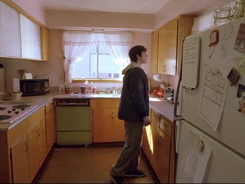 Movie still from “Elephant” (2003), directed by Gus Van Sant – A man standing in a kitchen next to a refrigerator; Wide shot, Low angle