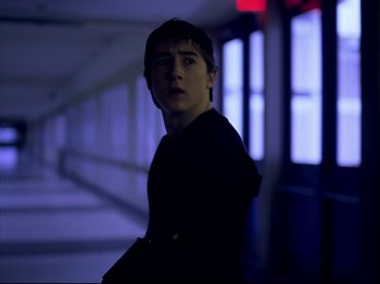 Movie still from “Elephant” (2003), directed by Gus Van Sant – A young man standing in a dark hallway; Medium shot, Over the shoulder angle