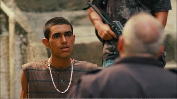 Movie still from “Elite Squad 2: The Enemy Within” (2010), directed by José Padilha – A young man with a chain around his neck and holding a gun; Medium shot, Over the shoulder angle