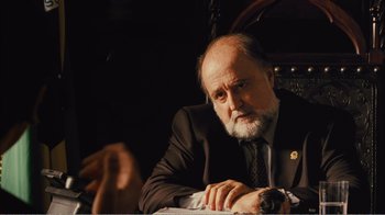 Movie still from “Elite Squad 2: The Enemy Within” (2010), directed by José Padilha – An older man wearing a suit and tie sitting at a table; Close Up shot, Over the shoulder angle
