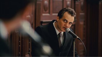 Movie still from “Elite Squad 2: The Enemy Within” (2010), directed by José Padilha – A man wearing a suit and tie holding a microphone in front of him; Close Up shot, Over the shoulder angle