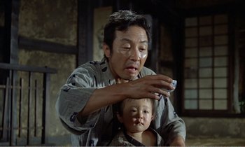 Movie still from “Empire of Passion” (1978), directed by Nagisa Ôshima – An older man and a young boy are sitting in front of a building; Medium shot, Low angle