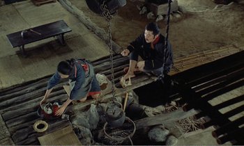 Movie still from “Empire of Passion” (1978), directed by Nagisa Ôshima – Two people sitting on a wooden platform near a fire pit; Wide shot, High angle
