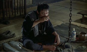 Movie still from “Empire of Passion” (1978), directed by Nagisa Ôshima – A man sitting on the ground eating something off of a spoon; Medium shot, High angle