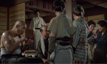 Movie still from “Empire of Passion” (1978), directed by Nagisa Ôshima – A group of people standing around a table; Medium shot, Over the shoulder angle