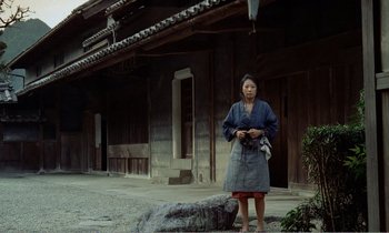 Movie still from “Empire of Passion” (1978), directed by Nagisa Ôshima – An older woman standing in front of a building; Wide shot, Low angle