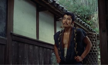 Movie still from “Empire of Passion” (1978), directed by Nagisa Ôshima – A man with a beard and a black shirt is standing in front of a building; Medium shot, Low angle