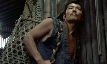 Movie still from “Empire of Passion” (1978), directed by Nagisa Ôshima – A man with a beard is standing next to a wooden structure; Medium shot, Low angle