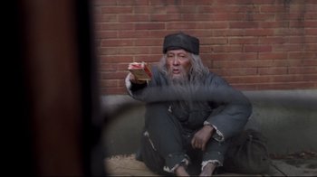 Movie still from “Empire of the Sun” (1987), directed by Steven Spielberg – An old man sitting on the ground holding a book; Medium shot, High angle
