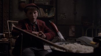 Movie still from “Empire of the Sun” (1987), directed by Steven Spielberg – A young boy sitting in front of a pan of food; Medium shot, Low angle