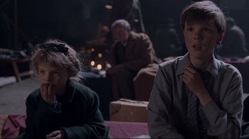 Movie still from “Empire of the Sun” (1987), directed by Steven Spielberg – Two people sitting on a couch in a dark room; Medium shot, Over the shoulder angle