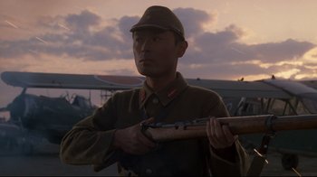 Movie still from “Empire of the Sun” (1987), directed by Steven Spielberg – A man in a uniform holding a rifle in front of an airplane; Close Up shot, Low angle