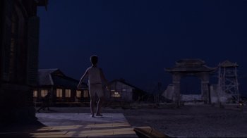 Movie still from “Empire of the Sun” (1987), directed by Steven Spielberg – A person standing on a sidewalk at night; Extreme Wide shot, High angle