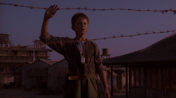 Movie still from “Empire of the Sun” (1987), directed by Steven Spielberg – A young man standing next to a barbed wire fence; Medium shot, Low angle