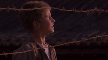 Movie still from “Empire of the Sun” (1987), directed by Steven Spielberg – A young man standing in front of a barbed wire fence; Close Up shot, Low angle