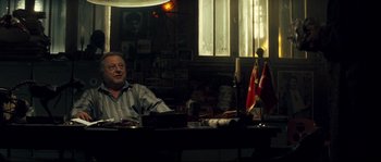Movie still from “Empire of the Wolves” (2005), directed by Chris Nahon – A man sitting at a table in a room; Medium shot, Over the shoulder angle