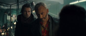 Movie still from “Empire of the Wolves” (2005), directed by Chris Nahon – A man in a leather jacket standing next to another man in a red shirt; Close Up shot, Over the shoulder angle